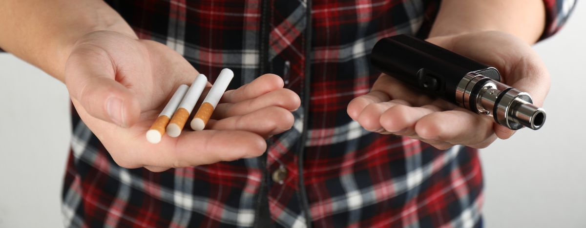 A person holds a vape in one hand and cigarettes in the other